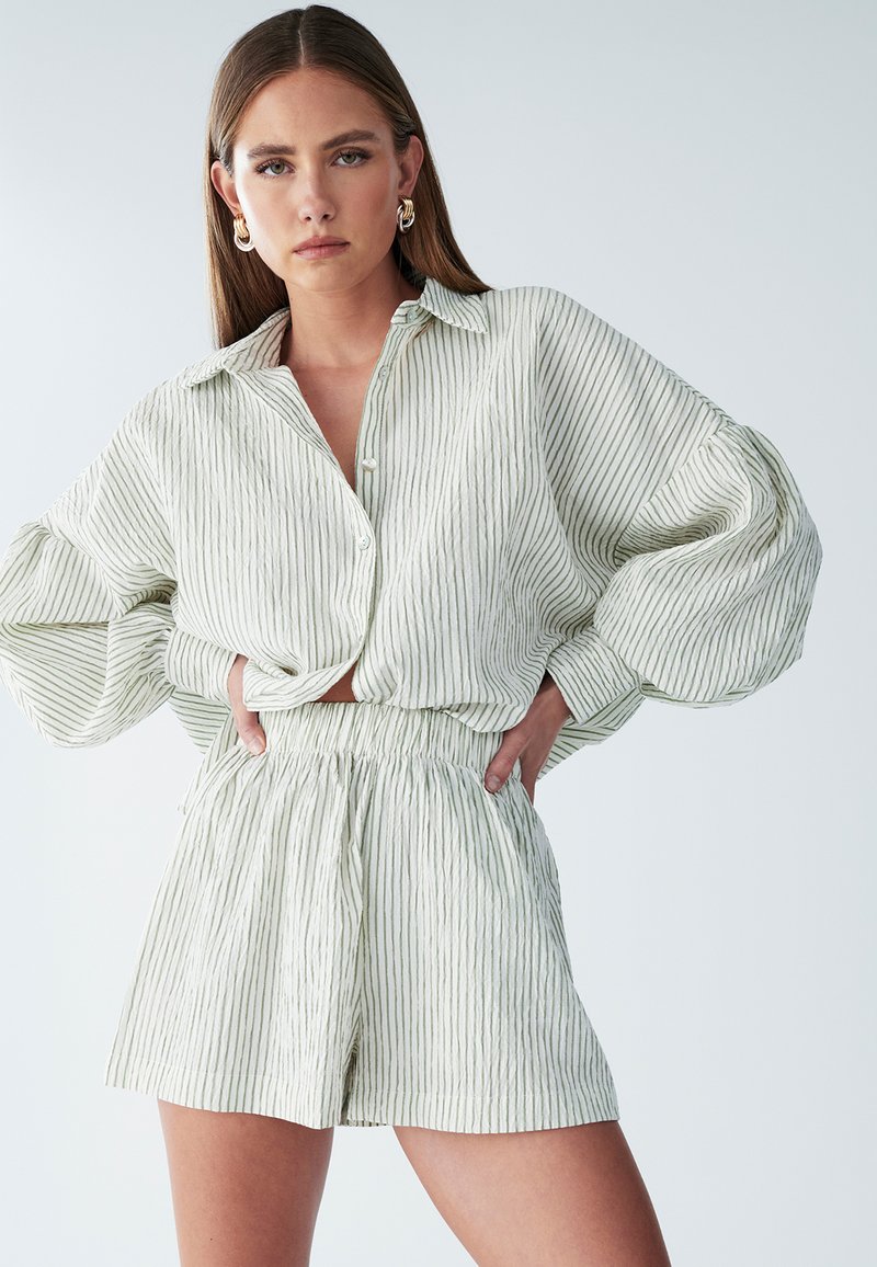 Žena pózujúca s rukami v bok, oblečená v voľnej bielej košeli s zelenými pruhmi a zodpovedajúcich krátkych nohaviciach s vysokým pásom.