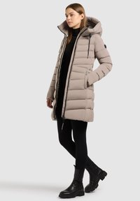 Beige pufferjack met een capuchon, voorzien van een ritssluiting aan de voorkant, zijzakken en een doorgestikt ontwerp. Gecombineerd met zwarte leggings en enkellaarzen.