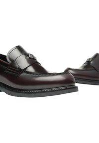 Un paio di mocassini in pelle marrone scuro con un emblema metallico decorativo sulle cinghie, dotati di suole in gomma nere e cuciture dettagliate.