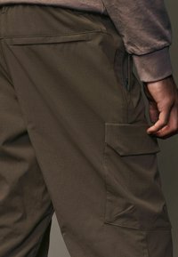 Pantalones cargo verde oliva con tela texturizada, bolsillo lateral con tapa y un bolsillo adicional con cremallera cerca de la cintura.