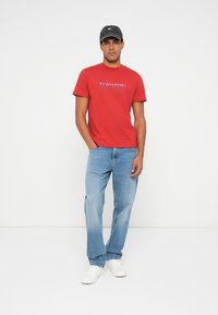 Camiseta de algodón roja con un logo azul, combinada con jeans de un azul claro y zapatillas blancas. El modelo lleva una gorra oscura. Diseño y ajuste simples.