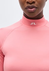 Κοντινό πλάνο ενός ατόμου που φοράει εφαρμοστή ροζ μπλούζα με ψηλό λαιμό και μικρό λευκό γεωμετρικό λογότυπο στο γιακά.