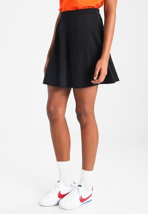 Personne portant une jupe noire en forme de trapèze, des baskets blanches Nike avec le logo Swoosh rouge, des chaussettes blanches et un haut orange, debout devant un fond blanc.
