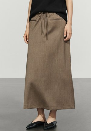 Kvinde iført en lang brun nederdel med snøre i taljen og to slidserlommer, kombineret med sorte slip-on sko og en sort top.