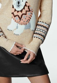 Pull beige confortable avec un motif d'ours polaire, des manches à motifs et des poignets côtelés. Associé à une jupe noire en simili cuir. Matière texturée et chaude.