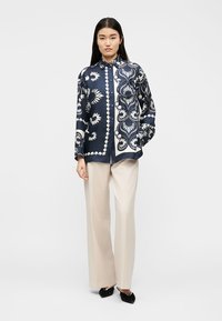 Γυναίκα που φοράει πουκάμισο με σχέδιο σε μπλε και λευκό χρώμα, μπεζ παντελόνι φαρδιάς γραμμής και μαύρα μυτερά παπούτσια, στέκεται με το ένα χέρι στην τσέπη.