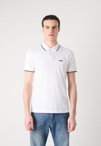 Biela polo košeľa s čiernymi akcentmi na golieri a rukávoch, s malým vyšívaným logom. Nosená s modrými džínami.