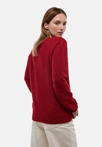 Pull en tricot rouge avec col rond, manches longues et poignets côtelés. Texture douce et coupe décontractée, présenté sur un fond neutre.