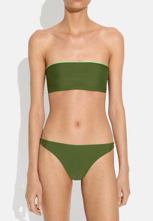 Kobieta ubrana w gładki oliwkowo-zielony, bezramiączkowy top od bikini i pasujące majtki bikini na białym tle.