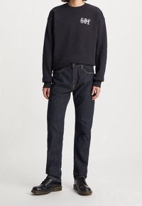 Personne portant un sweat-shirt noir avec le texte "501", un jean bleu foncé et des chaussures en cuir noires à lacets, debout devant un fond uni.