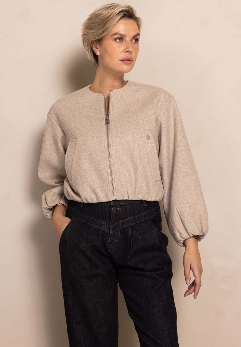 Light beige zip-up jacket with a round collar and balloon sleeves, paired with dark denim high-waisted pants featuring gold stitching details.