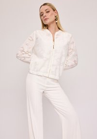 Biała kurtka bomberka z fakturowanej tkaniny i długimi, przezroczystymi rękawami. Posiada zamek błyskawiczny z przodu i elastyczny pas. W zestawie z białymi spodniami.