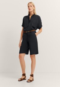 Dunkelmarineblaues Kurzarmhemd mit Knopfleiste, kombiniert mit knielangen Shorts. Ein brauner Gürtel betont die Taille und wird von braunen Knöchel-Sandalen ergänzt.