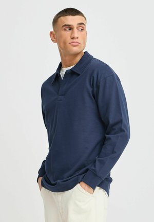 Marineblaues Langarm-Polohemd mit Knopfkragen, aus glattem Stoff gefertigt. Es hat einen flachen Saum und eine lockere Passform. Kombiniert mit hellen Hosen.