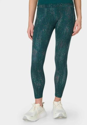Benen met groene legging met patroon en witte sportschoenen zichtbaar van taille tot voeten op witte achtergrond.