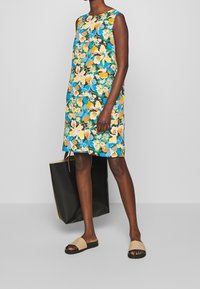 Vestido floral en tonos azules, amarillos y verdes; diseño sin mangas, hasta la rodilla, combinado con sandalias beige y un gran bolso negro.
