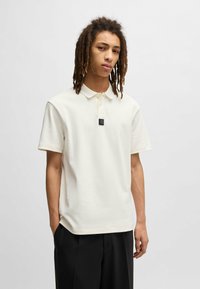 Cremfarbendes Poloshirt mit kurzen Ärmeln, lockerer Passform, zwei Knöpfen und einem schwarzen rechteckigen Logo auf der Brust. Aus glattem Stoff gefertigt.