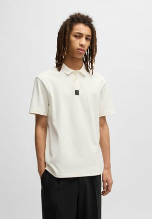 Cremfarbendes Poloshirt mit kurzen Ärmeln, lockerer Passform, zwei Knöpfen und einem schwarzen rechteckigen Logo auf der Brust. Aus glattem Stoff gefertigt.