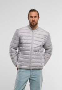 Lys grå polstret jakke i bomberstil, med lynlås fortil, to sidelommer og ribbet krave. Glat, blank overflade.