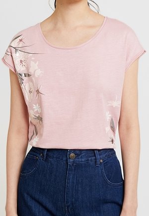 Frau trägt ein hellrosa kurzärmeliges Shirt mit Blumen- und Insektenmustern an den Seiten, kombiniert mit hoch taillierter dunkelblauer Jeans.