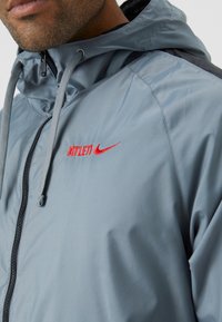 Lys grå vindjakke med hætte, prydet med røde "ATHLETI" og Nike-logoer, sidepaneler og lynlås. Glat tekstur.