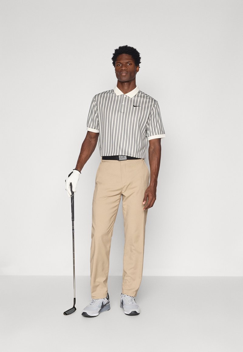 Polo con colletto a righe verticali grigie e bianche, pantaloni beig, cintura nera, guanto da golf bianco e scarpe da ginnastica argento/grigie.