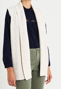 Stickad ärmlös väst i krämfärg med texturerad, öppen stickning och ribbad nederkant. Matchad med en mörk skjorta och olivgröna byxor.