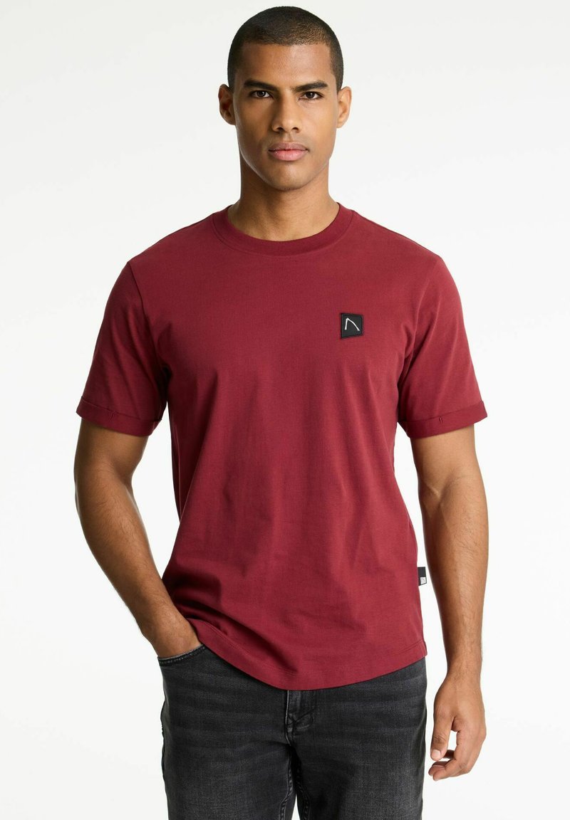 Rotes Baumwoll-T-Shirt mit kurzen Ärmeln, runder Ausschnitthals und einem kleinen schwarzen Logo-Patch auf der Brust. Getragen mit dunklen Jeans.
