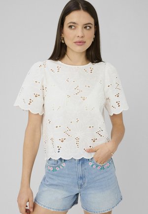 Femme portant un chemisier blanc à manches courtes en dentelle œil-de-perdrix et un short en jean bleu clair avec broderies florales, posant avec une main dans la poche.