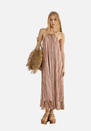 Mujer con un vestido marrón sin mangas hasta el suelo que sostiene un gran bolso de paja tejido y lleva sandalias con plataforma.