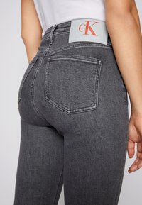 Jeans de mezclilla gris con cintura alta, cinco bolsillos y una etiqueta de cuero con el logo "Calvin Klein" en naranja en la parte trasera de la cintura.