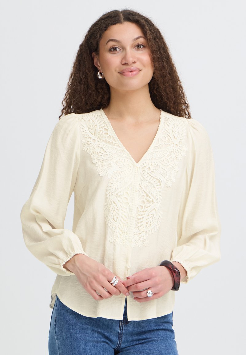 Jeune femme aux longs cheveux bouclés portant un chemisier crème avec des détails en dentelle et un jean bleu, les mains jointes devant elle, souriant doucement.