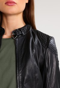 Black leather jacket with a high collar, featuring quilted shoulder accents and a zipper front. Worn over a green top. Smooth texture.