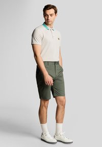 Ung mand iført beige polo med teal krave, grønne shorts, hvide sokker og hvide sneakers, står med hænderne i lommerne på en grå baggrund.