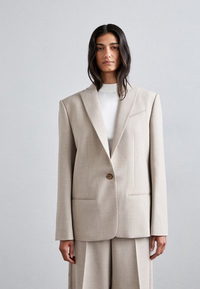 Beiger, taillierter Blazer mit ausgeprägten Schultern, einem Ein-Knopf-Verschluss und dezenten Steppdetails, kombiniert mit einem weißen Rollkragenpullover und weiten Hosen.