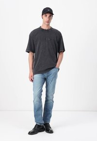Gray oversized t-shirt with raw hem, logo detail, paired with light blue jeans and black shoes. Plain background, model stands upright.