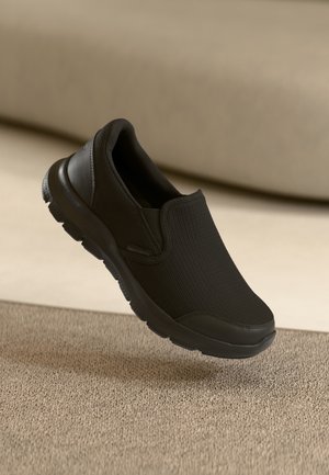 Chaussure Skechers noire à enfiler avec dessus texturé et semelle en caoutchouc épaisse, montrée flottant au-dessus d'une surface beige texturée.