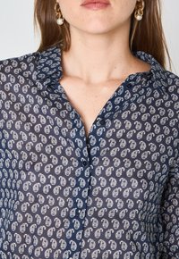 Blusa a fantasia blu navy con colletto, chiusura a bottoni e intricate decorazioni a paisley bianche su un tessuto trasparente.
