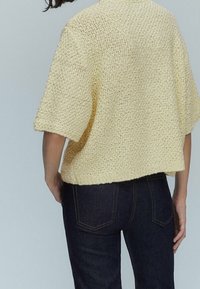 Żółty sweter z dzianiny o luźnym fasonie, krótkich rękawach i teksturowanym wzorze, w połączeniu z ciemnymi dżinsami.