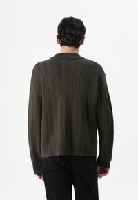 Άνδρας με κοντά σκούρα μαλλιά που φοράει μια σκούρα ελιά, ριμπ μπλούζα και μαύρο παντελόνι, στέκεται με τα πίσω προς μια λευκή προοπτική.