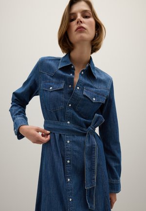 Denim jurk met lange mouwen, twee borstzakken en een strikriem. Heeft knopen aan de voorkant en een diepblauwe kleur met een licht vervaagde textuur.