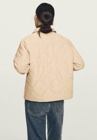 Beżowa pikowana kurtka z płaskim kołnierzem, ozdobiona falistymi przeszyciami; w połączeniu z luźnymi niebieskimi dżinsami, widok od tyłu.