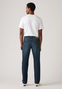 Muž stojaci chrbtom, oblečený v bielom tričku, tmavomodrých džínsoch s vreckami na zadnej strane a bielych teniskách na jednoduchom pozadí.