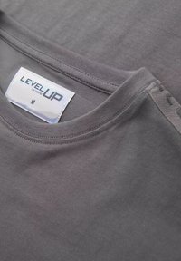 Šedé bavlněné tričko s kulatým výstřihem. Obsahuje prošívaný lem a štítek s nápisem "LEVEL UP" v tučných, reflexních písmenech. Velikost M.