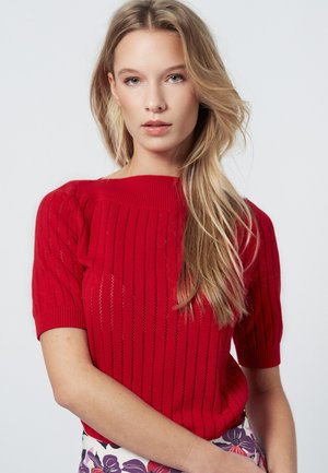 Rode gebreide trui met korte mouwen, voorzien van verticale strepen en een geribde halslijn. Het model draagt het met een rok met een bloemenpatroon.