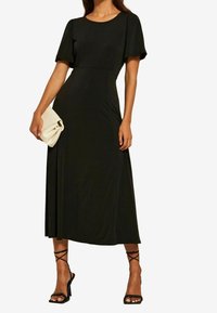 Musta midi-mekko sileästä kankaasta, jossa on lyhyet hihat, vyötä oleva vyötärö ja vajaalätyskä. Yhdistettynä kermanväriseen käsilaukkuun ja mustiin nauhakorkokenkiin.