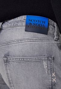 Jeans de denim gris claro con detalles desgastados, que presentan una etiqueta de cuero azul oscuro grabada con el logo "Scotch & Soda".