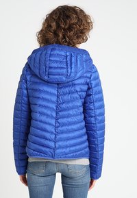 Blå pufferjakke med kviltede lodrette linjer, fastgjort hætte og ribbet krave. Kombineret med grå talje og afslappet denim.