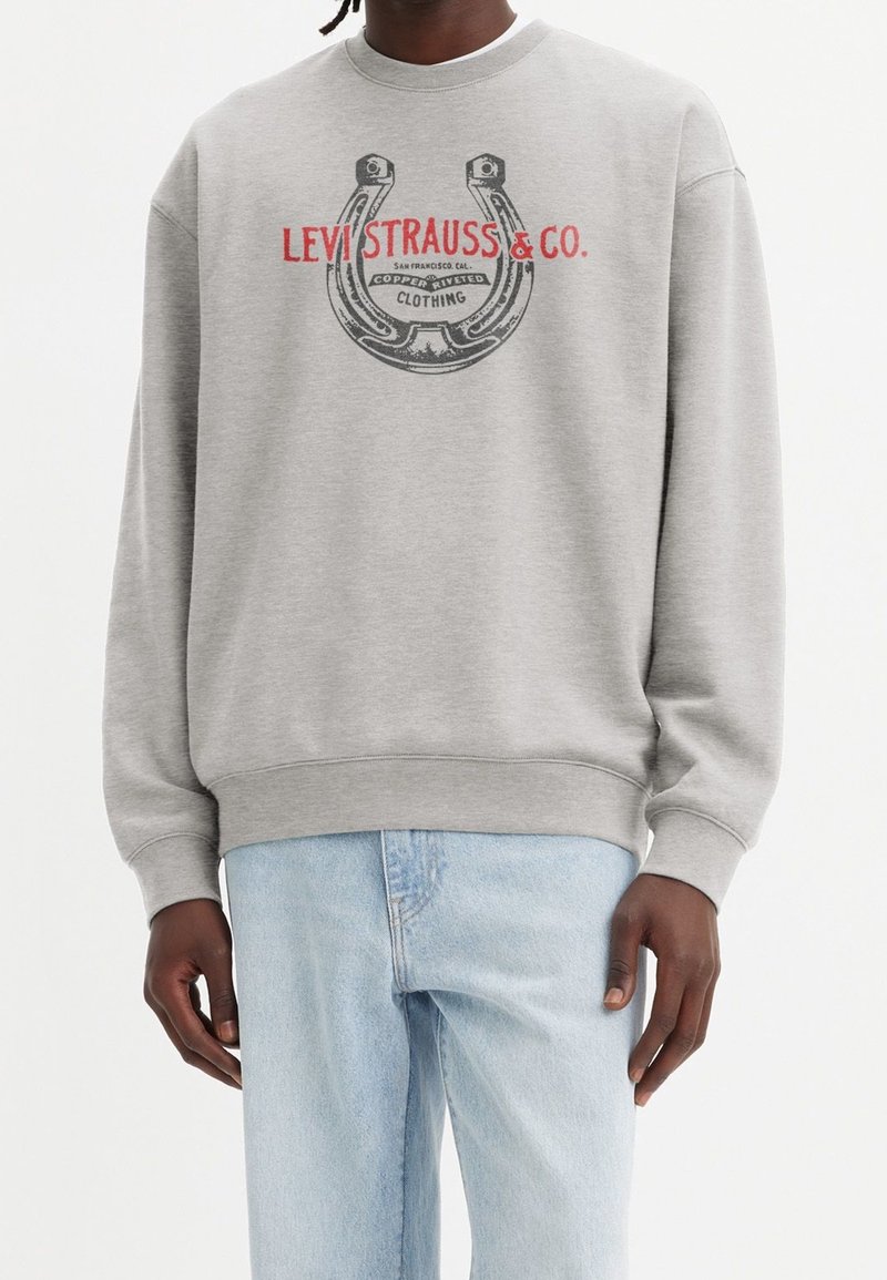 Sudadera gris hecha de algodón con cuello redondo, que presenta un logo de herradura impreso y texto en negro y rojo. Corte holgado con puños acanalados.