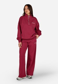 Burgundijas krāsas zip-svilpes krekls un atbilstošas bikses mīkstajā audumā, piedāvājot brīvu piegriezumu, logo detaļu un plašus kāju galiņus.
